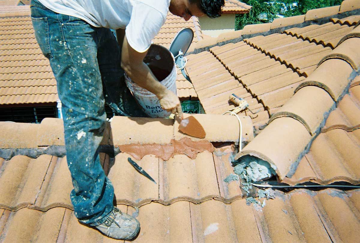 Worker repairing terracotta roof tiles on a rooftop with a hammer and safety harness