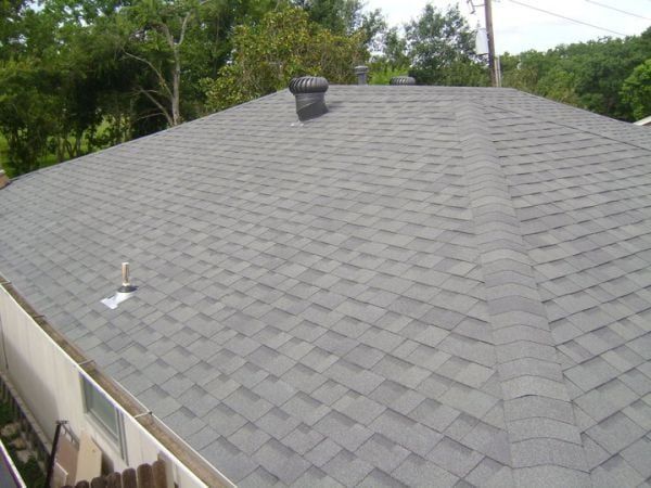 Gray shingled house roof with a vent pipe and trees in the background