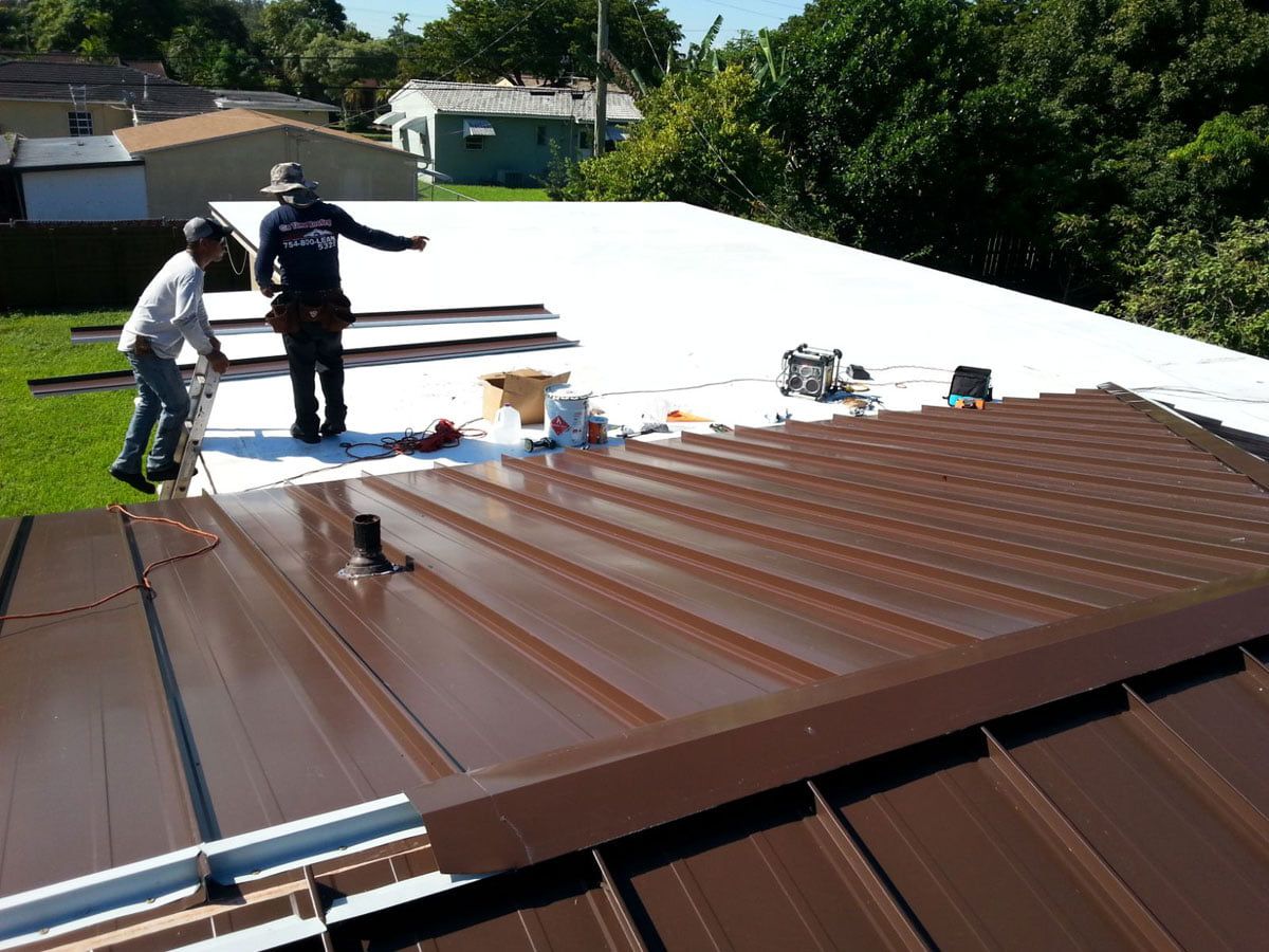Workers on a rooftop installing brown metal roofing beside a large white roof section.
