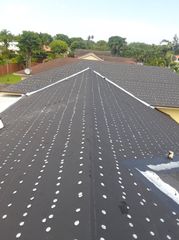 Roof under construction with dark underlayment and rows of white fasteners, trees in the background