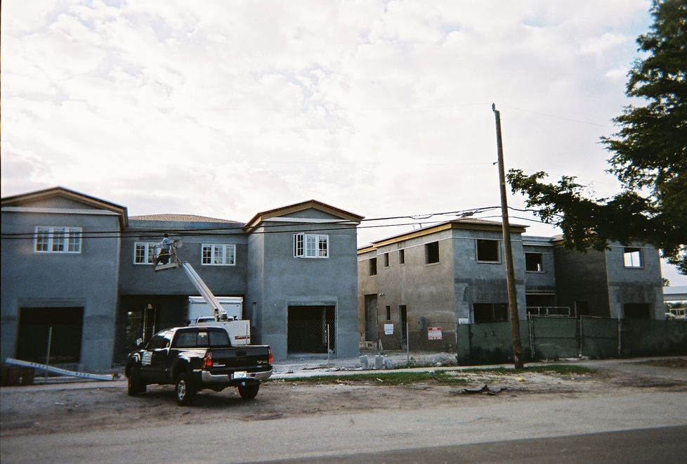 Row of unfinished gray houses along a dirt road, with a pickup truck parked in front.