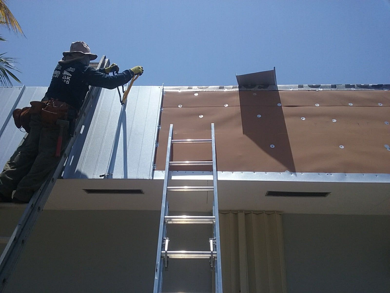 Worker on ladder installing brown siding on a building against a blue sky