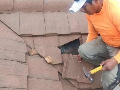 Worker repairing brown shingle roof with a hammer near a damaged section