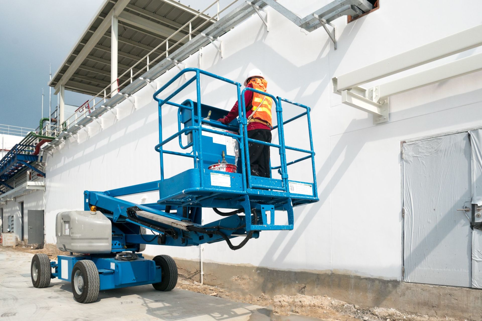 Un operaio su una piattaforma aerea blu dipinge un muro industriale bianco all'aperto.