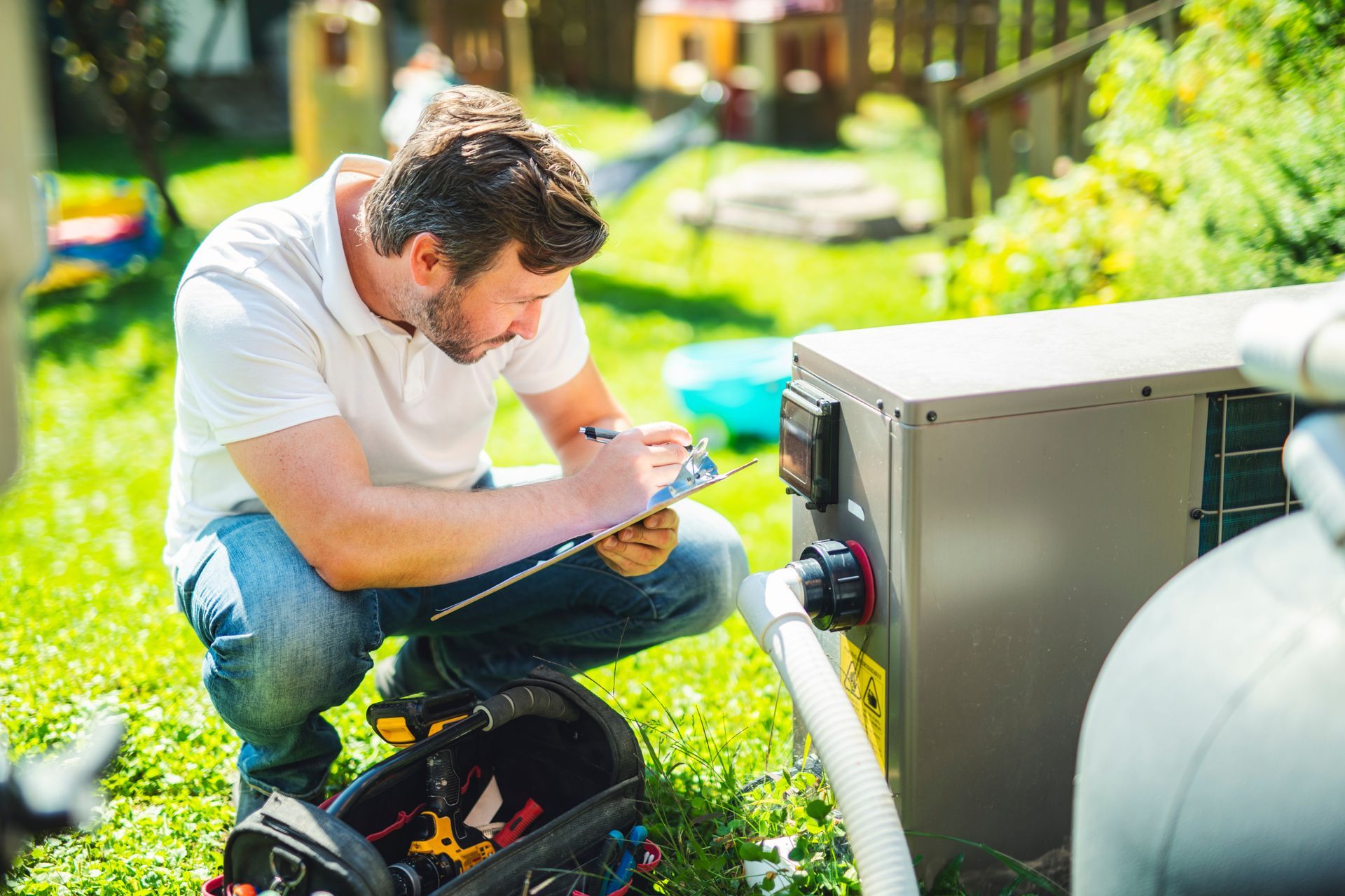 A man is kneeling down in the grass looking at a pool pump.