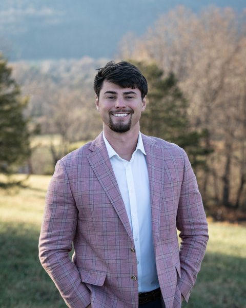 A smiling person in a light pink textured blazer and white dress shirt stands in a grassy field with trees in the back.