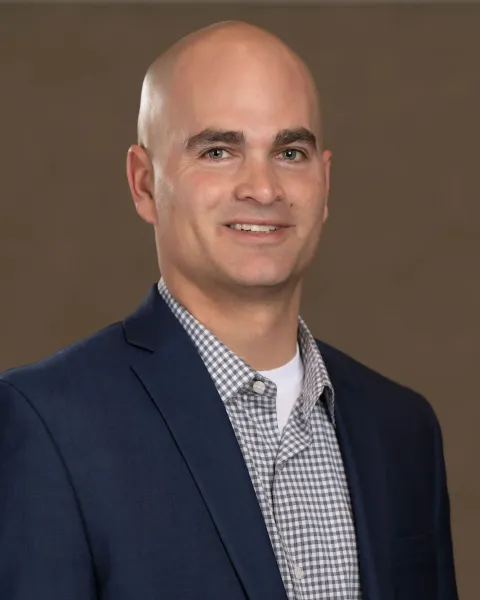 Headshot of a smiling person wearing a navy blazer and a grey patterned button-down shirt against a neutral background.