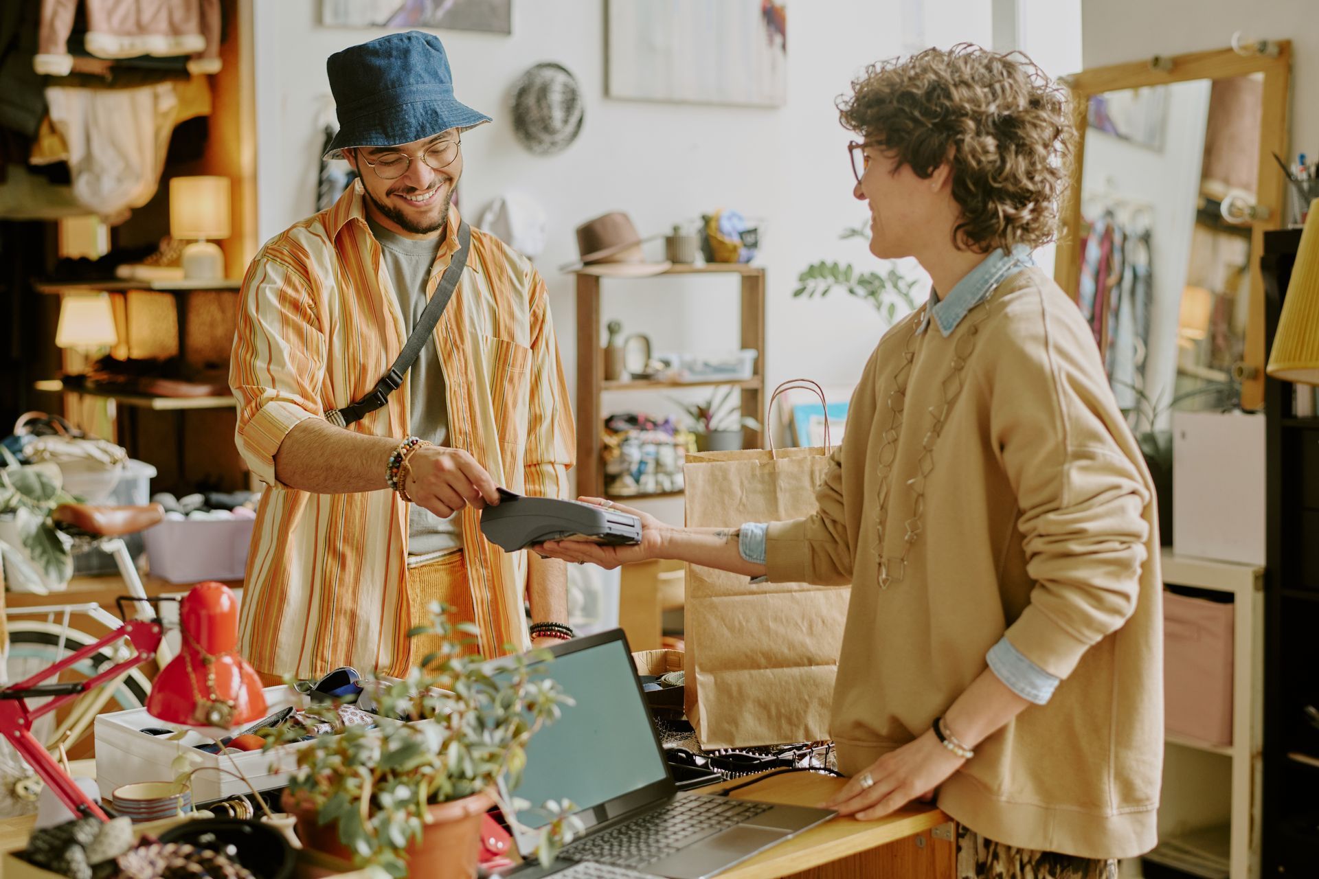 A customer pays at a shop counter, holding a payment device while the cashier looks on in a cluttered retail store.