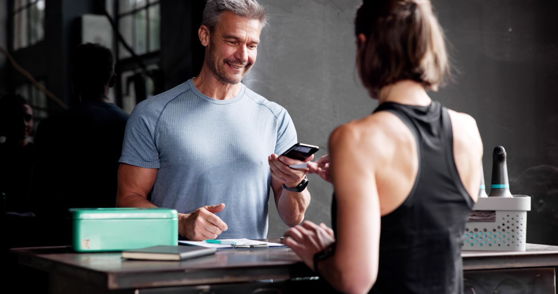 A smiling person in a blue athletic shirt holds a smartphone to scan a code at a gym front desk for an associate.