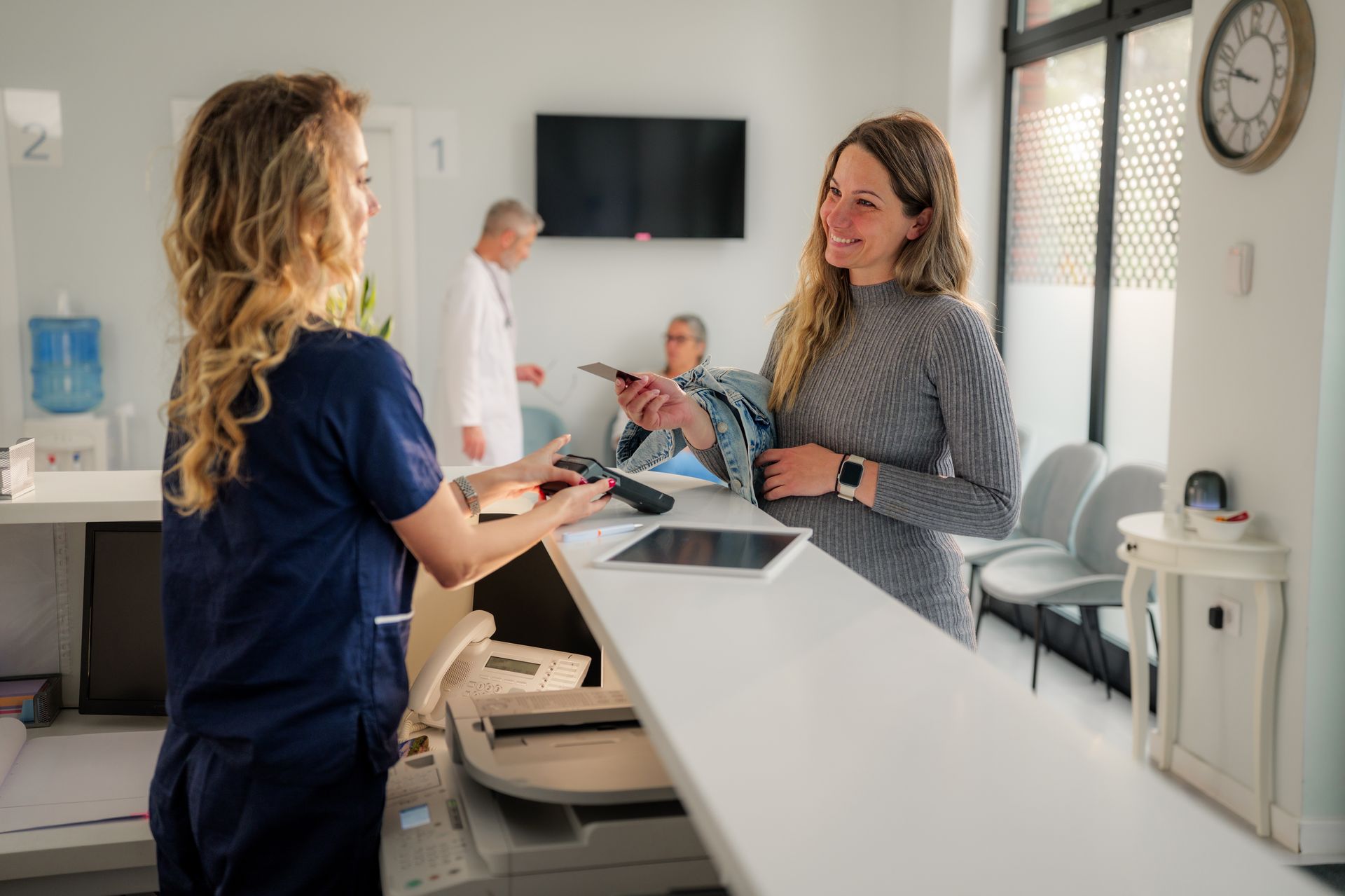 A patient hands a payment terminal to a receptionist at a medical office reception desk.