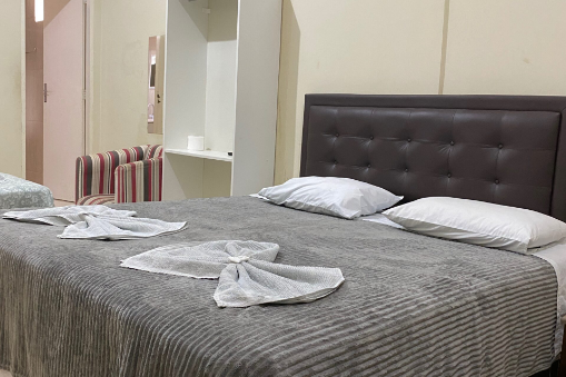 Quarto com colcha cinza, travesseiros brancos, toalhas dobradas e cabeceira marrom.