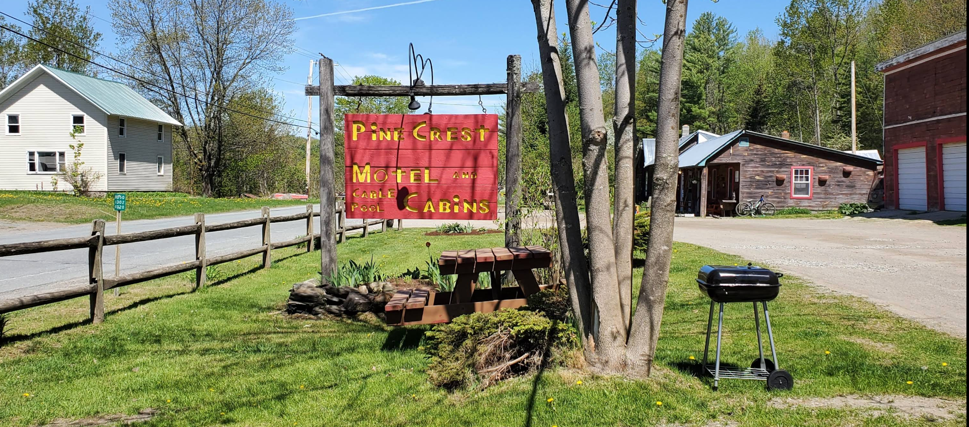 Red roadside sign by picnic table and grill on a grassy lot with trees and houses nearby