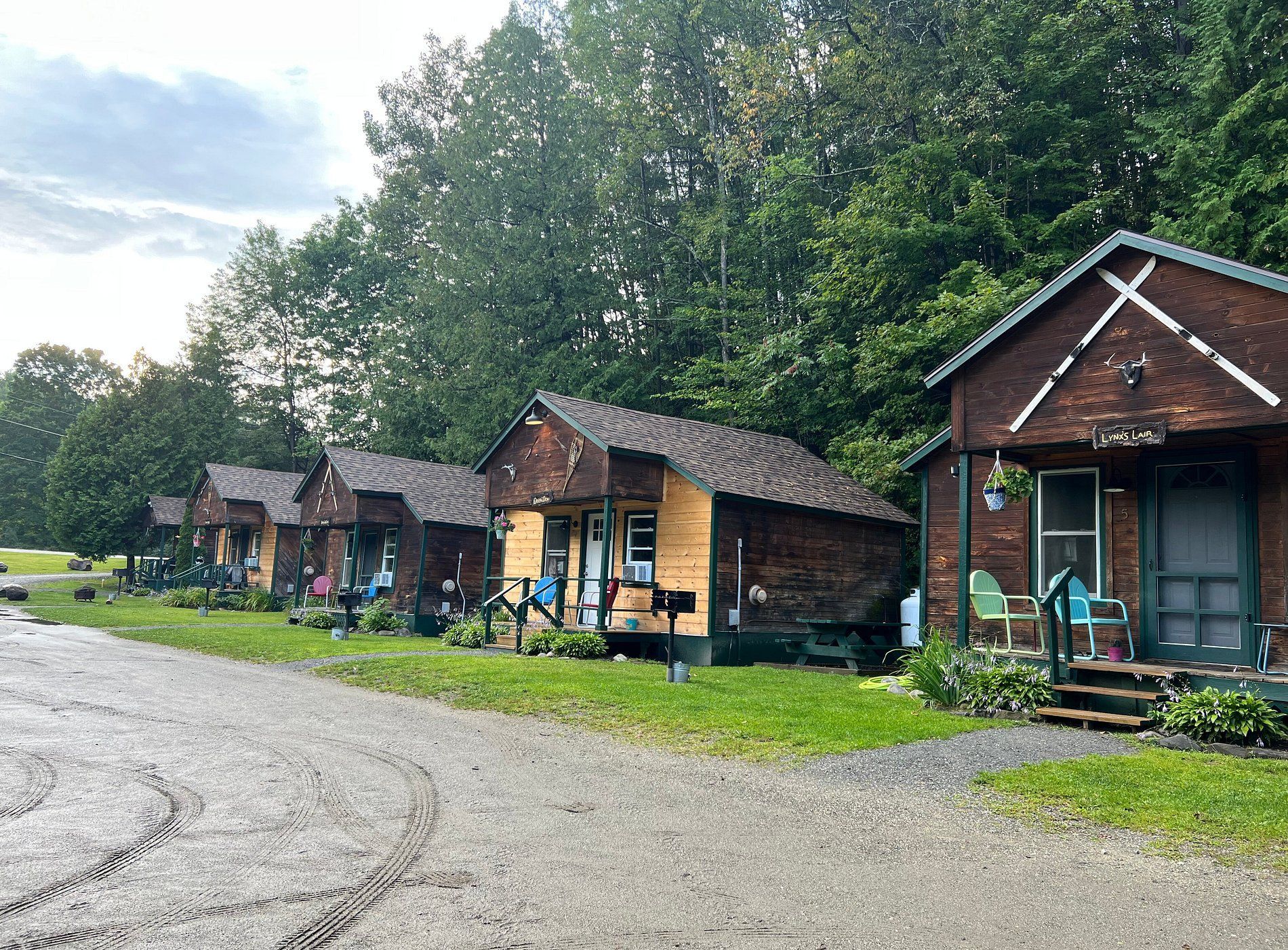 Rustic cabins line a gravel path beside a wooded area on a cloudy day.