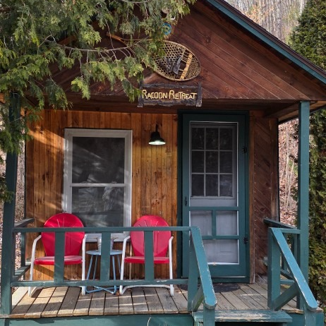 Small rustic cabin porch with two red chairs and a teal door under a wooden awning.