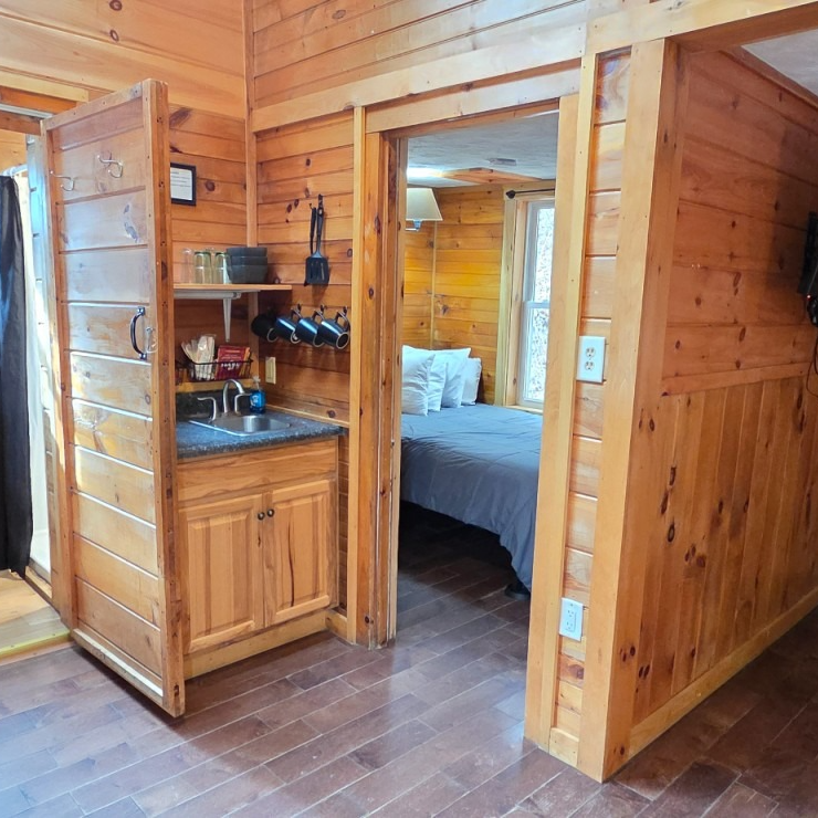 Wood-paneled cabin interior with a small kitchenette and doorway to a bedroom with a bed.