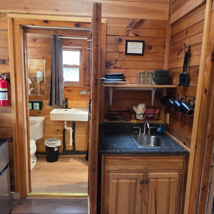Small rustic wood cabin kitchenette with sink, shelves, and a doorway to a bathroom with toilet and sink