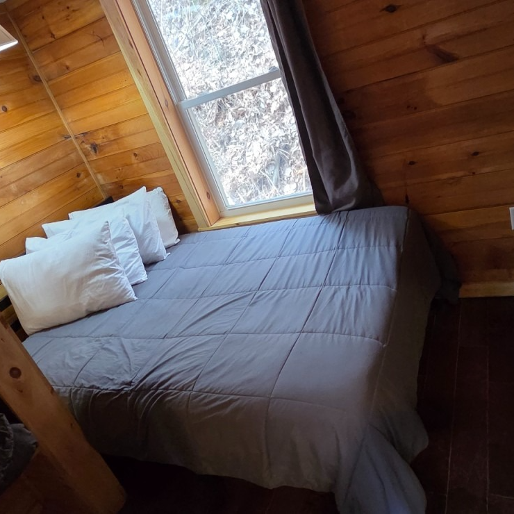 Wood-paneled cabin bedroom with a gray bedspread, white pillows, and a window beside the bed