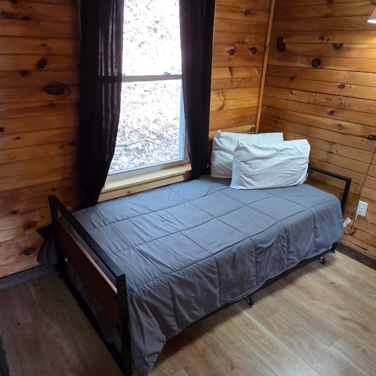 Single bed with gray bedding in a small wood-paneled room beside a window and dark curtains