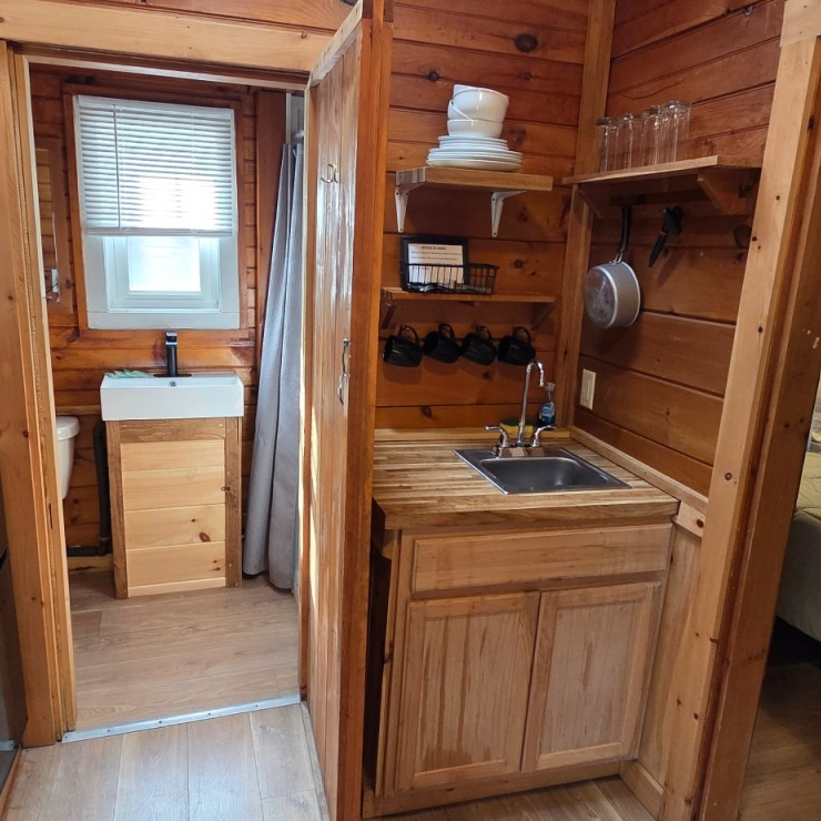 Rustic cabin bathroom with wood walls, small sink vanity, toilet, and open doorway to a bedroom.