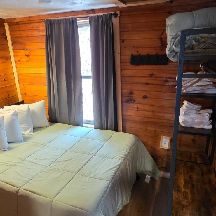 Cozy cabin bedroom with wooden walls, a bed with gray bedding, window curtains, and a bunk bed shelf.