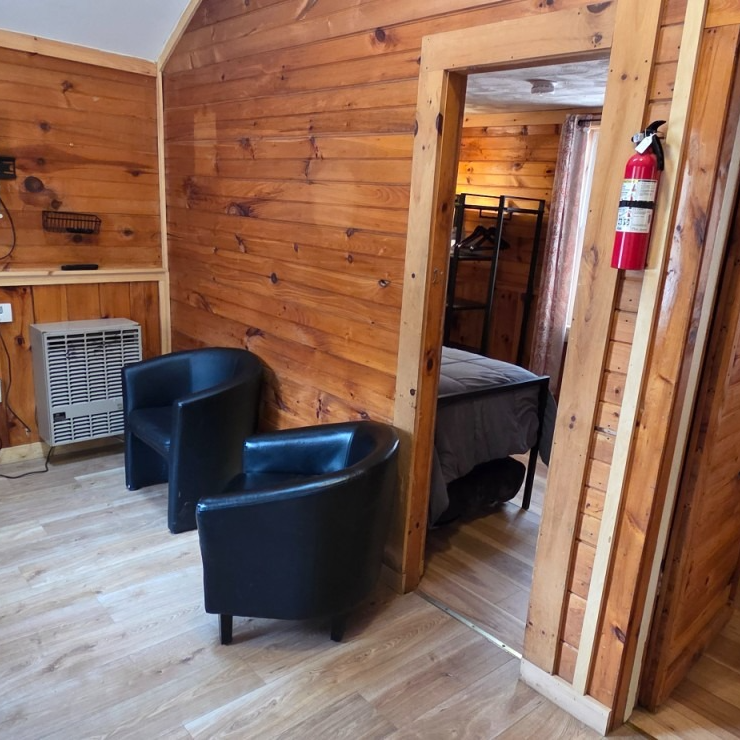 Cozy wood-paneled room with two black chairs, a small heater, and a doorway to a bedroom.