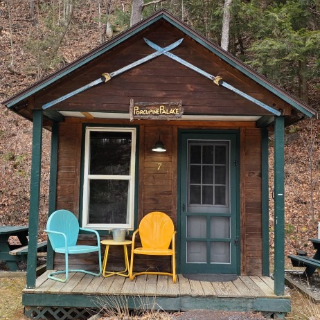 Small rustic cabin porch with turquoise and yellow chairs, a green door, and a “Rockin’ B” sign.