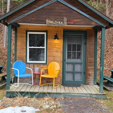 Small rustic cabin porch with blue and yellow chairs, pink table, and a green door under a lit porch lamp.