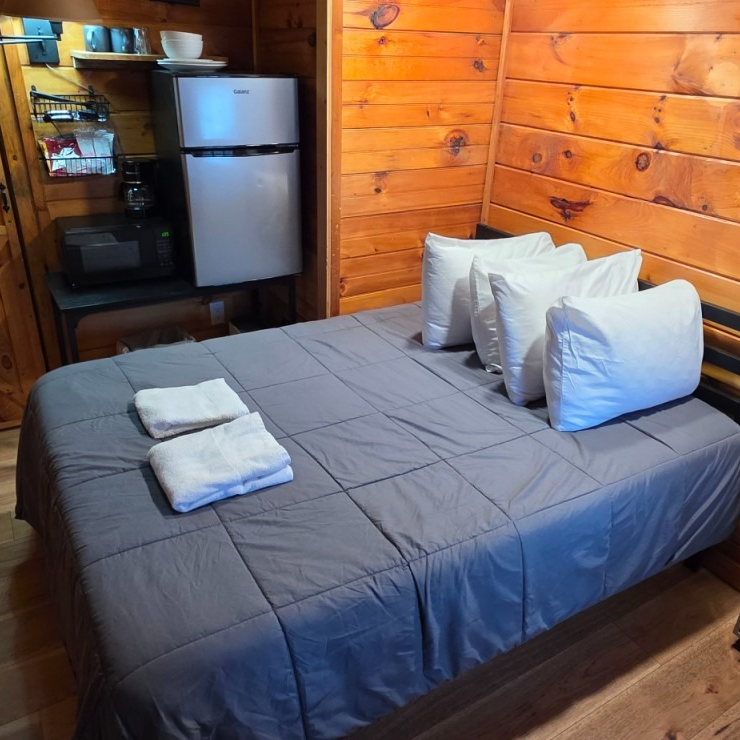 Cozy wooden cabin bedroom with a gray bedspread, white pillows, and a small kitchenette in the background