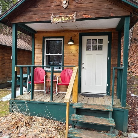 Small rustic cabin porch with two pink chairs, teal railings, and a white door.