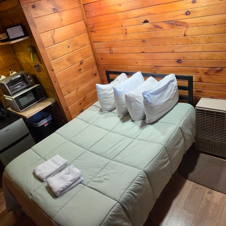 Small wood-paneled bedroom with a neatly made twin bed, folded towels, and a small kitchenette shelf.