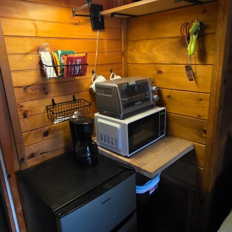 Small wood-paneled kitchenette with a mini fridge, microwave, coffee maker, toaster oven, and hanging bananas.
