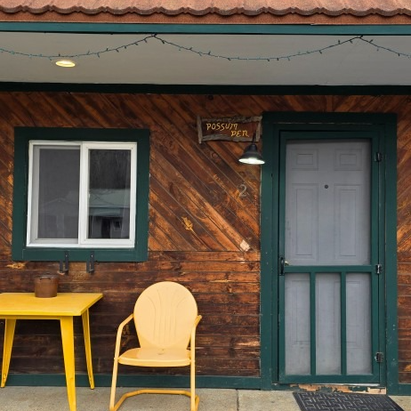 Rustic cabin porch with green-trimmed window and door, yellow table and chair, and a hanging light.