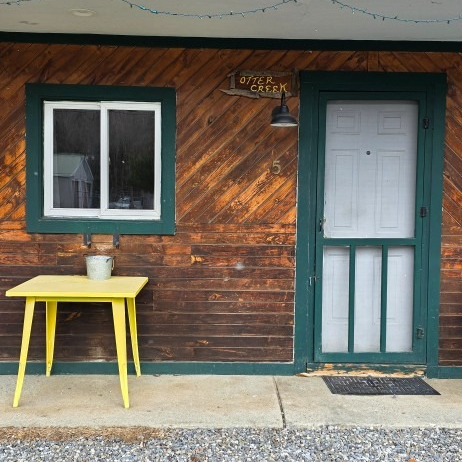 Rustic cabin porch with green-trimmed window and door, yellow table, and a bucket on the table