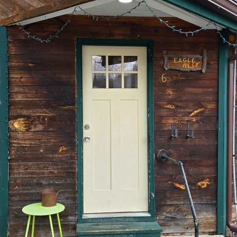 Weathered wooden cabin entrance with cream door, hanging lights, and a small green table with a pot.
