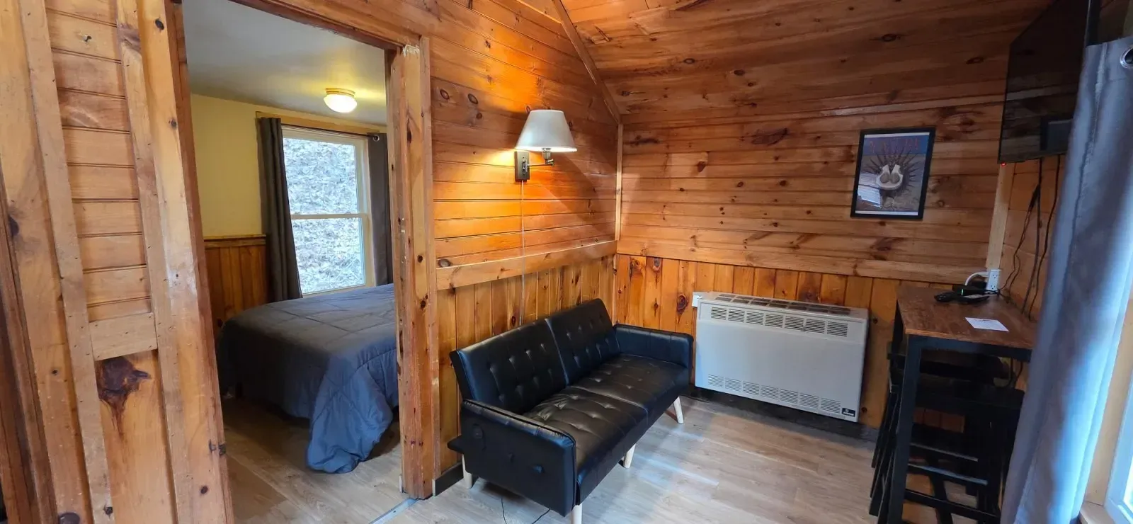 Cozy wooden cabin interior with a bed, black sofa, radiator, and window view