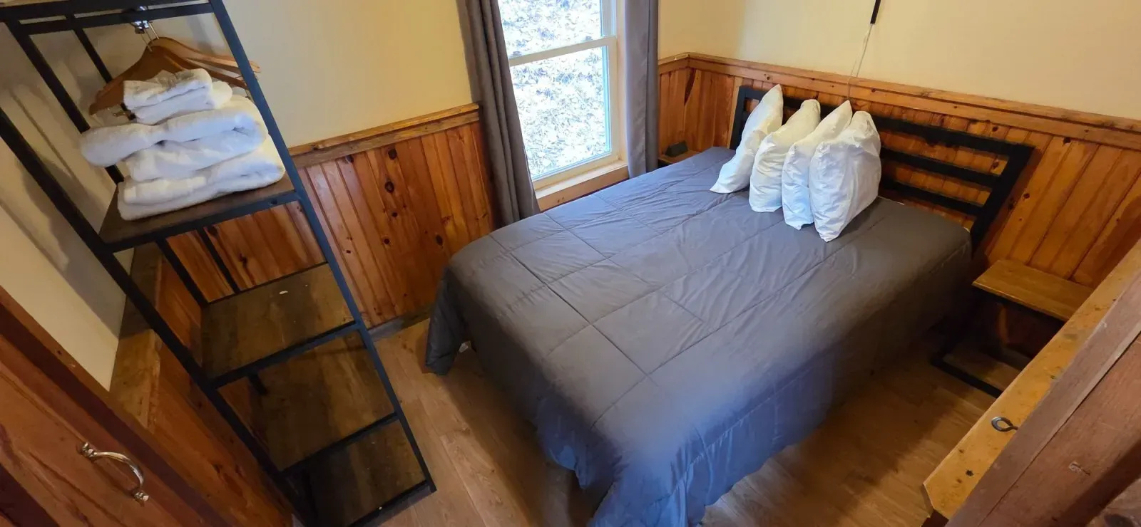 Cozy wood-paneled bedroom with a double bed, gray bedding, and a window letting in natural light.