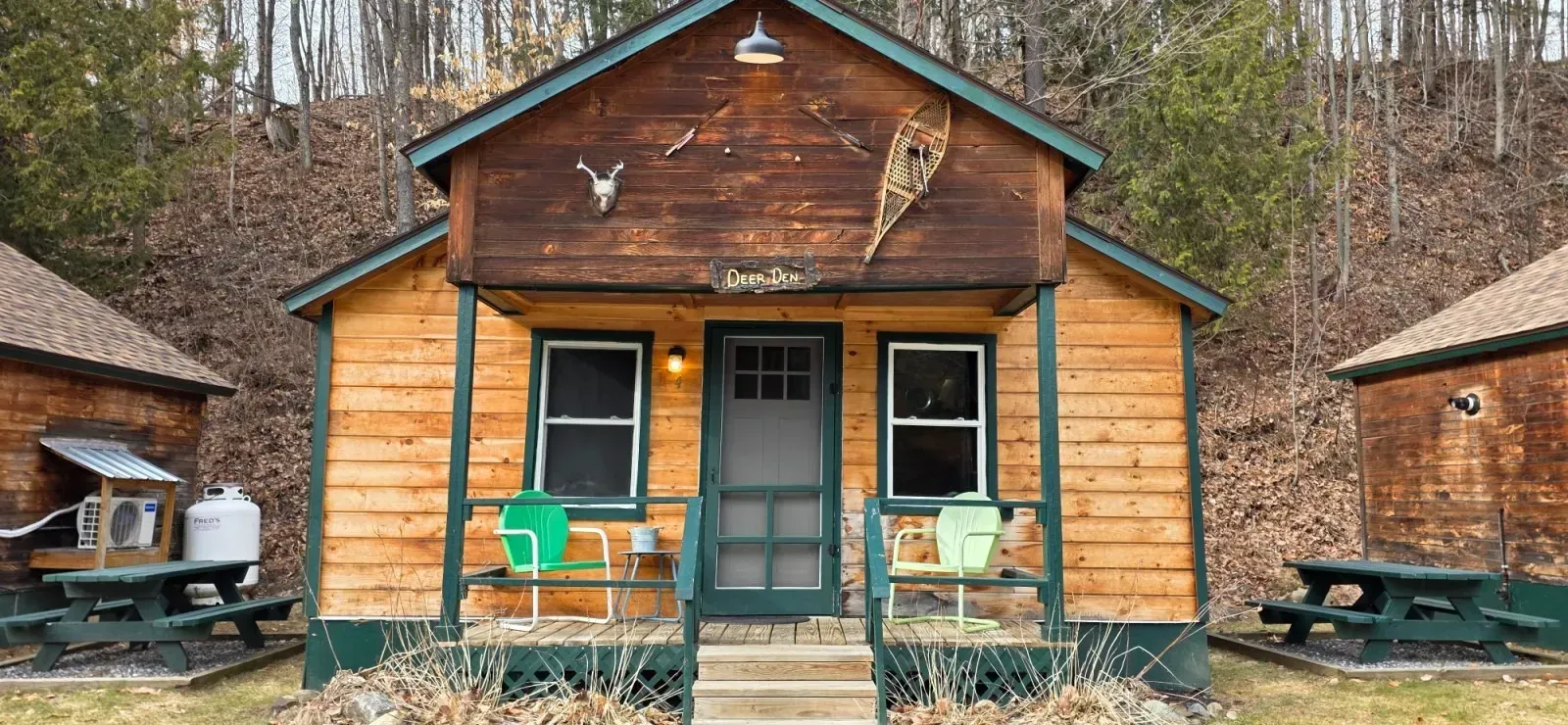 Small rustic cabin with green doors and two picnic tables in a wooded setting