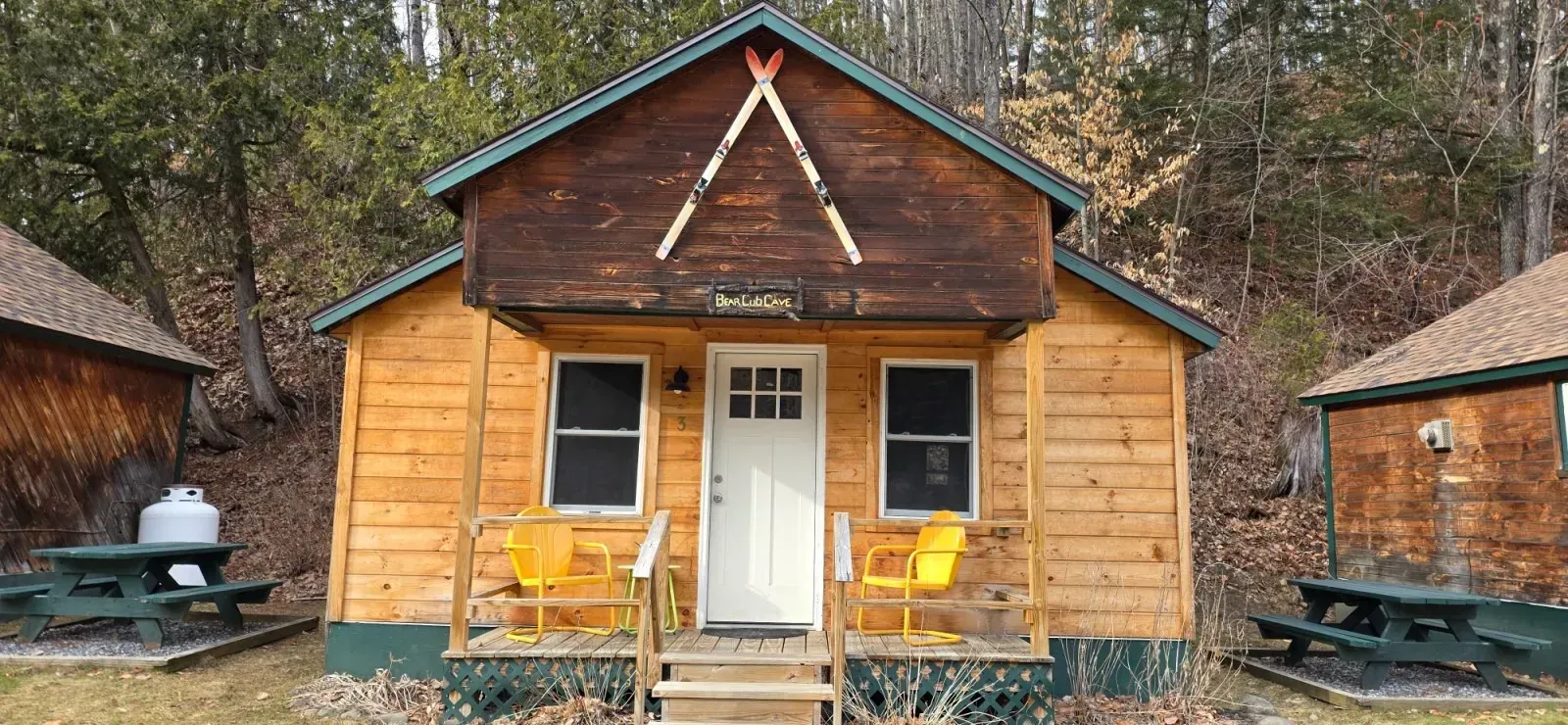 Small rustic cabin with a white door, yellow chairs on the porch, and picnic tables in a wooded setting