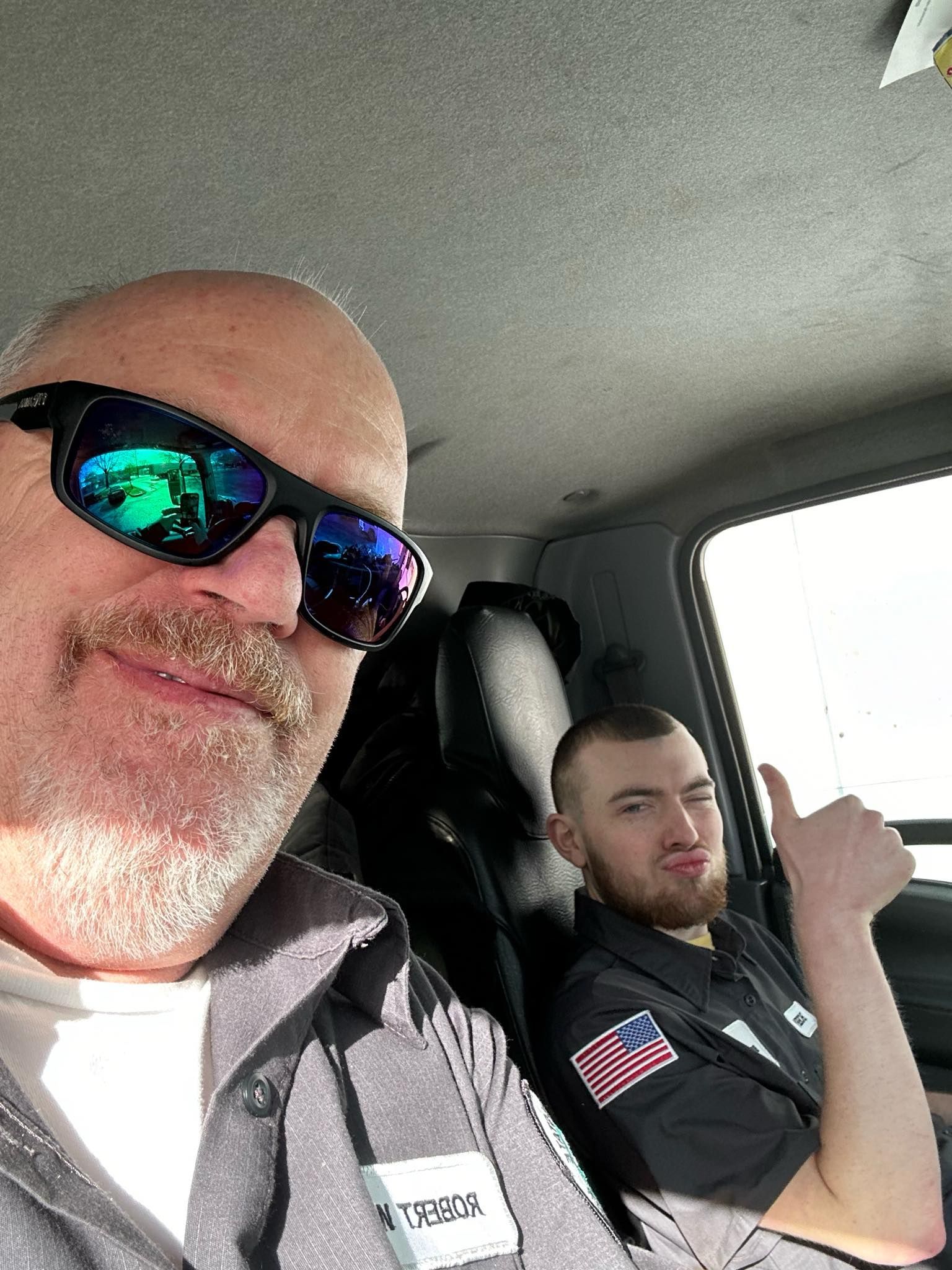 Two men sit in a vehicle; one wears sunglasses, the other gives a thumbs-up.