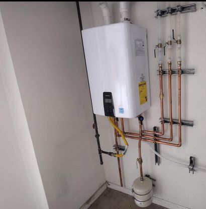 White wall-mounted water heater with copper pipes and a small floor tank in a utility room
