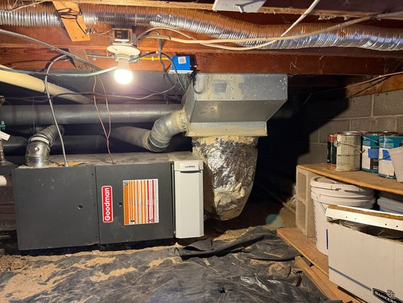 Dim basement with furnace, exposed ductwork, light bulb, and storage items under low wooden joists