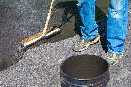 Person applying black sealant to asphalt with a squeegee near a bucket