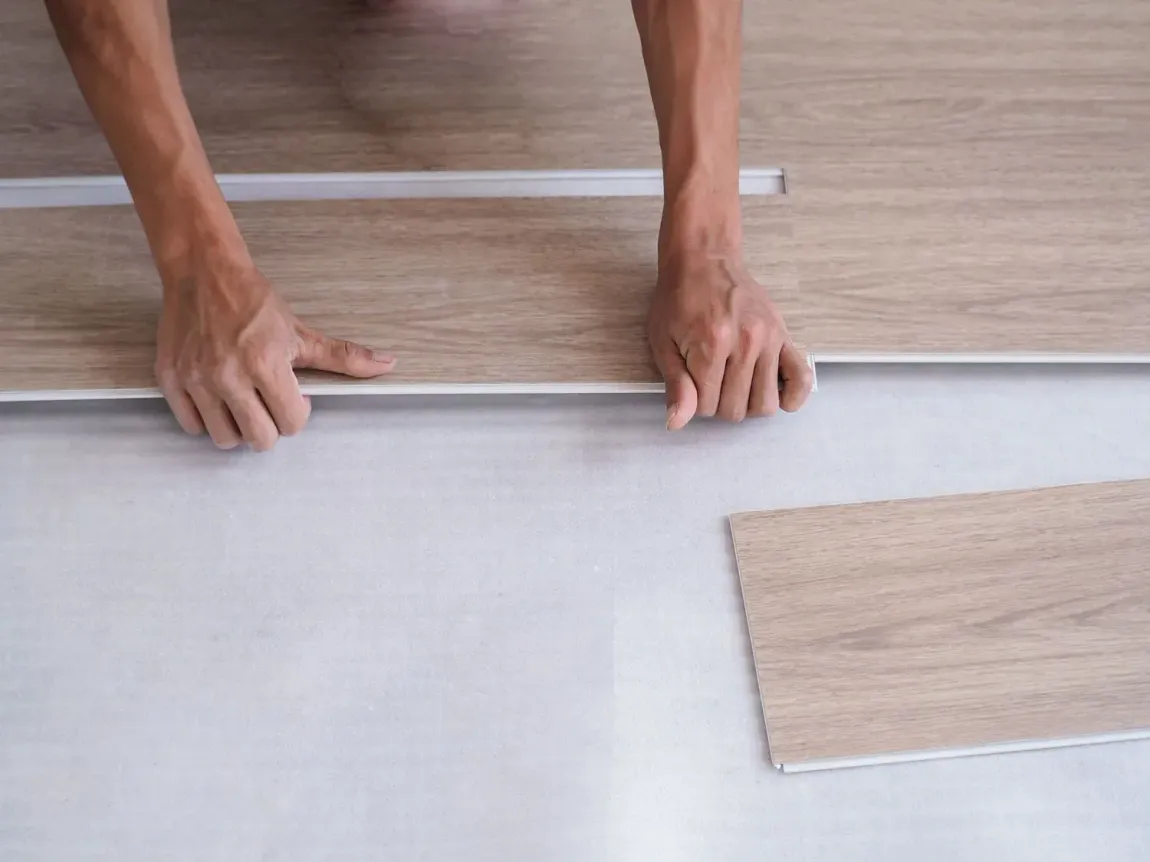 Hands laying a white underlayment beside light wood flooring planks on a floor.