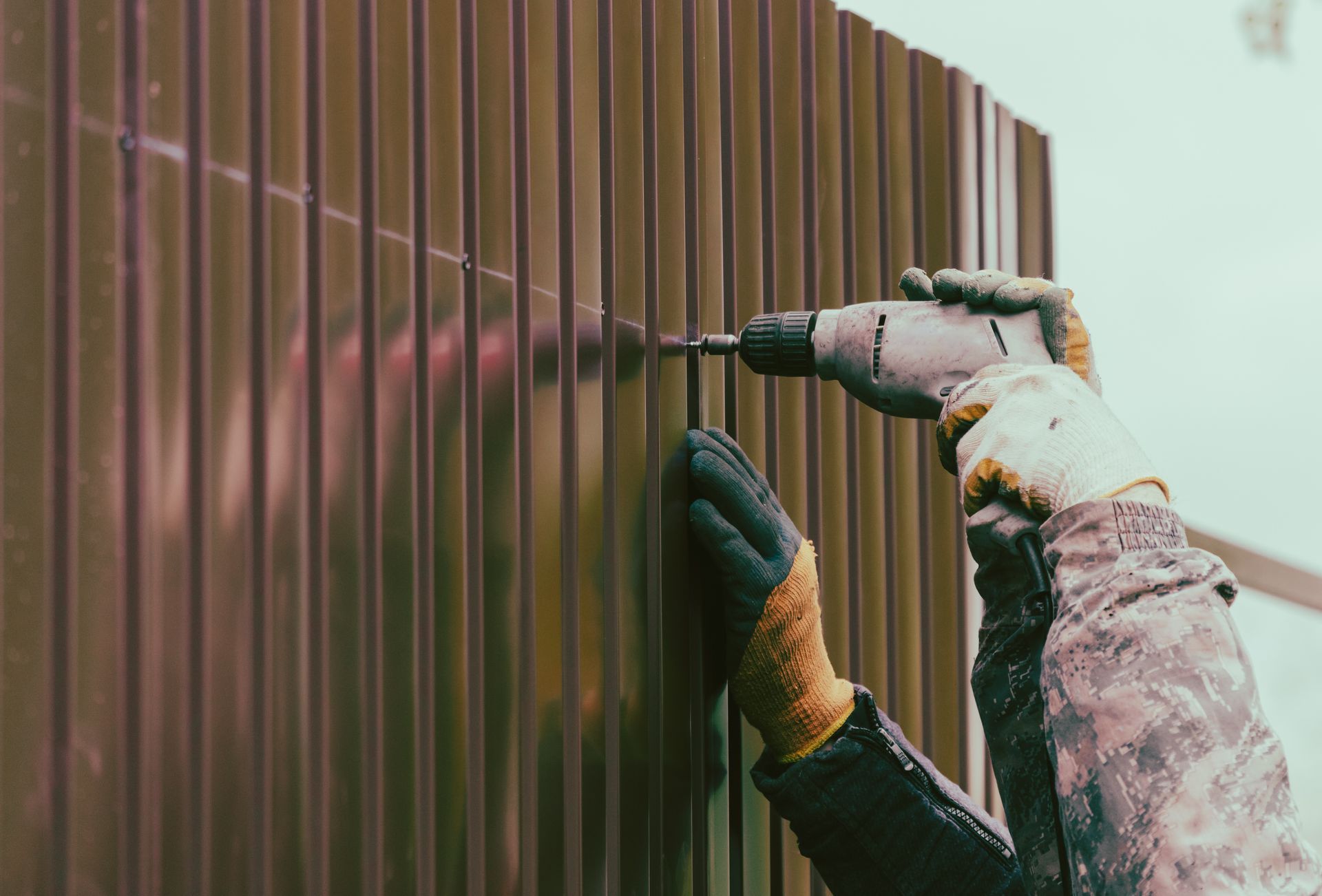 Gloved hands drill a brown metal fence panel with a power tool.