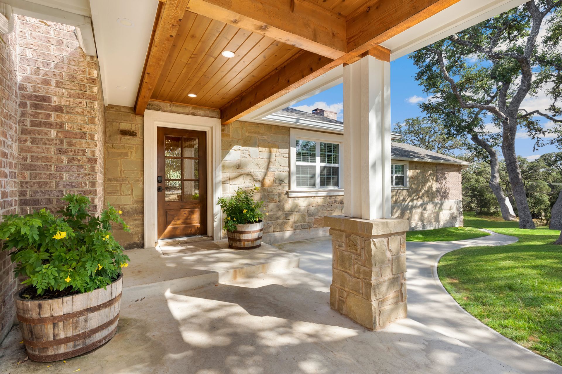 Covered front porch with wood ceiling, stone columns, potted plants, and a view of the yard.