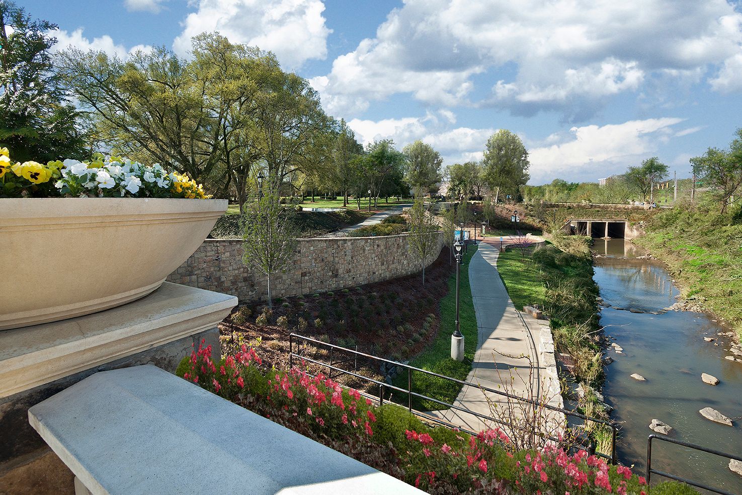 Park-like riverside promenade with stone wall, path, and flowering planters.