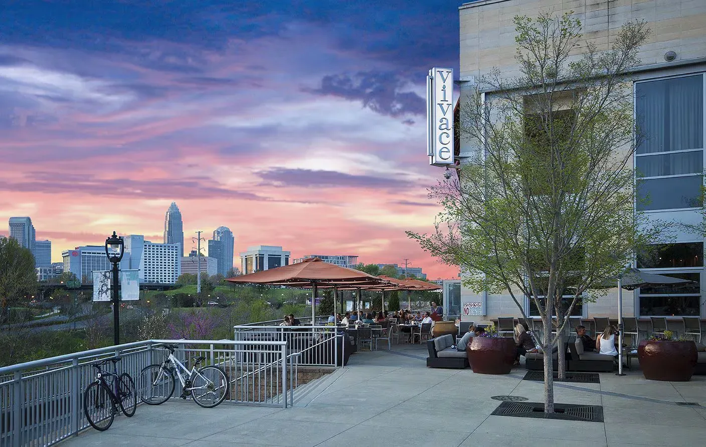 Restaurant patio overlooking city skyline at sunset. People dine at tables under orange umbrellas, bikes parked on railing.
