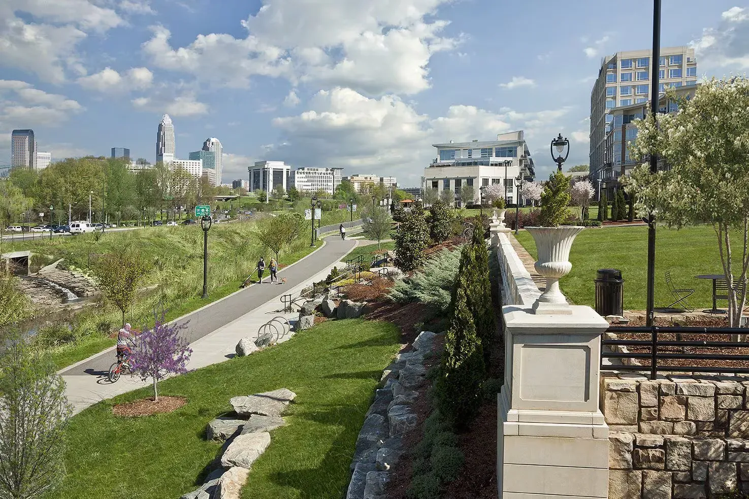 Park pathway with greenery, stone walls, and city skyline in the distance under a partly cloudy sky.