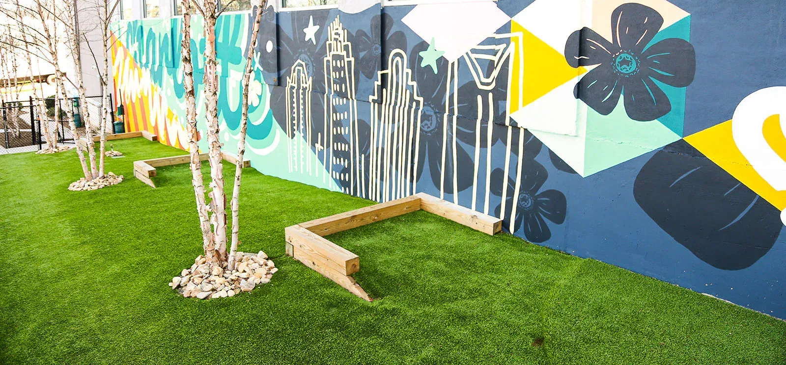 Outdoor community courtyard with a colorful mural wall, artificial grass, trees, and wooden seating.
