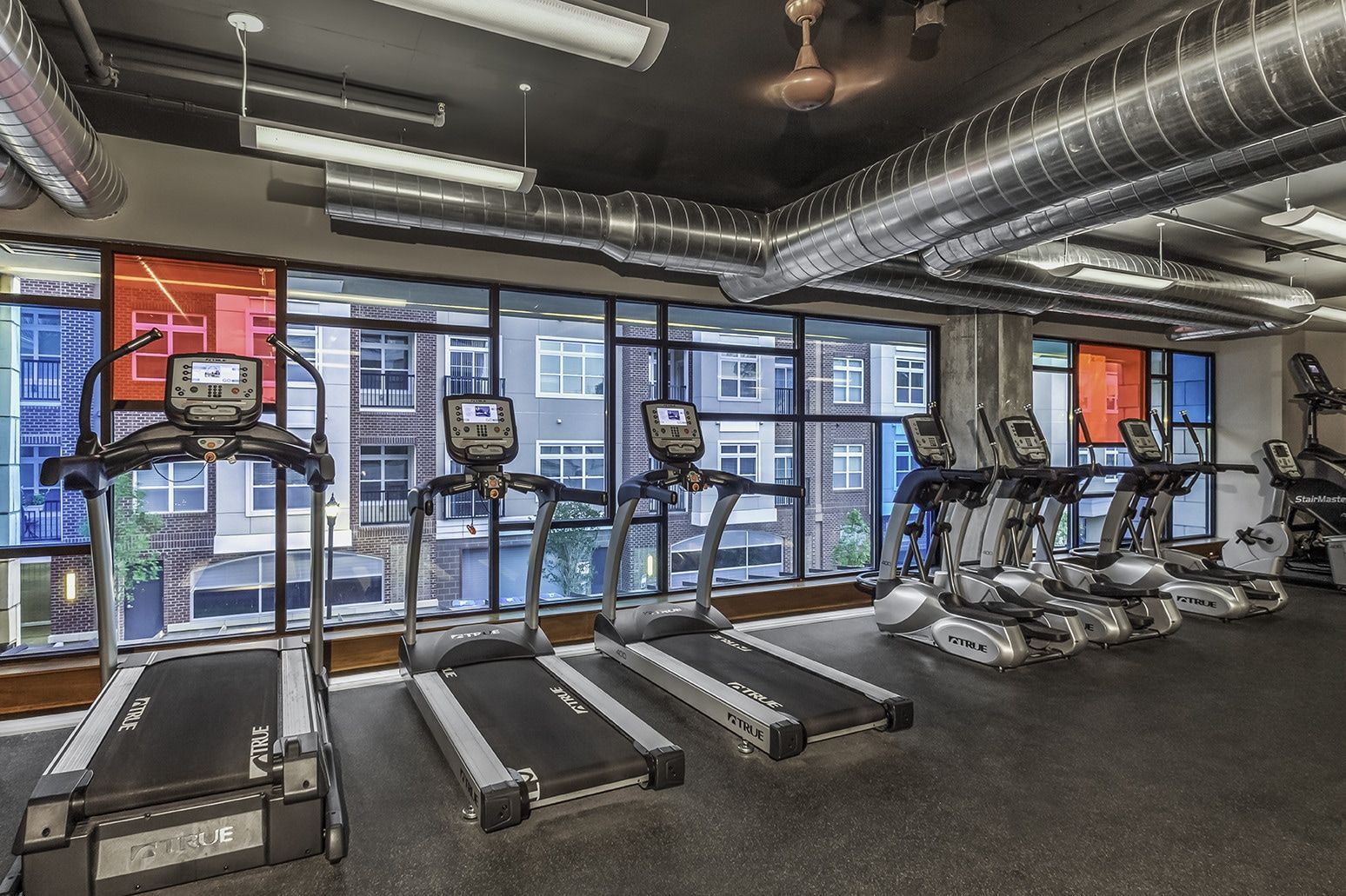 Gym interior with treadmills and elliptical machines, windows overlooking a building.